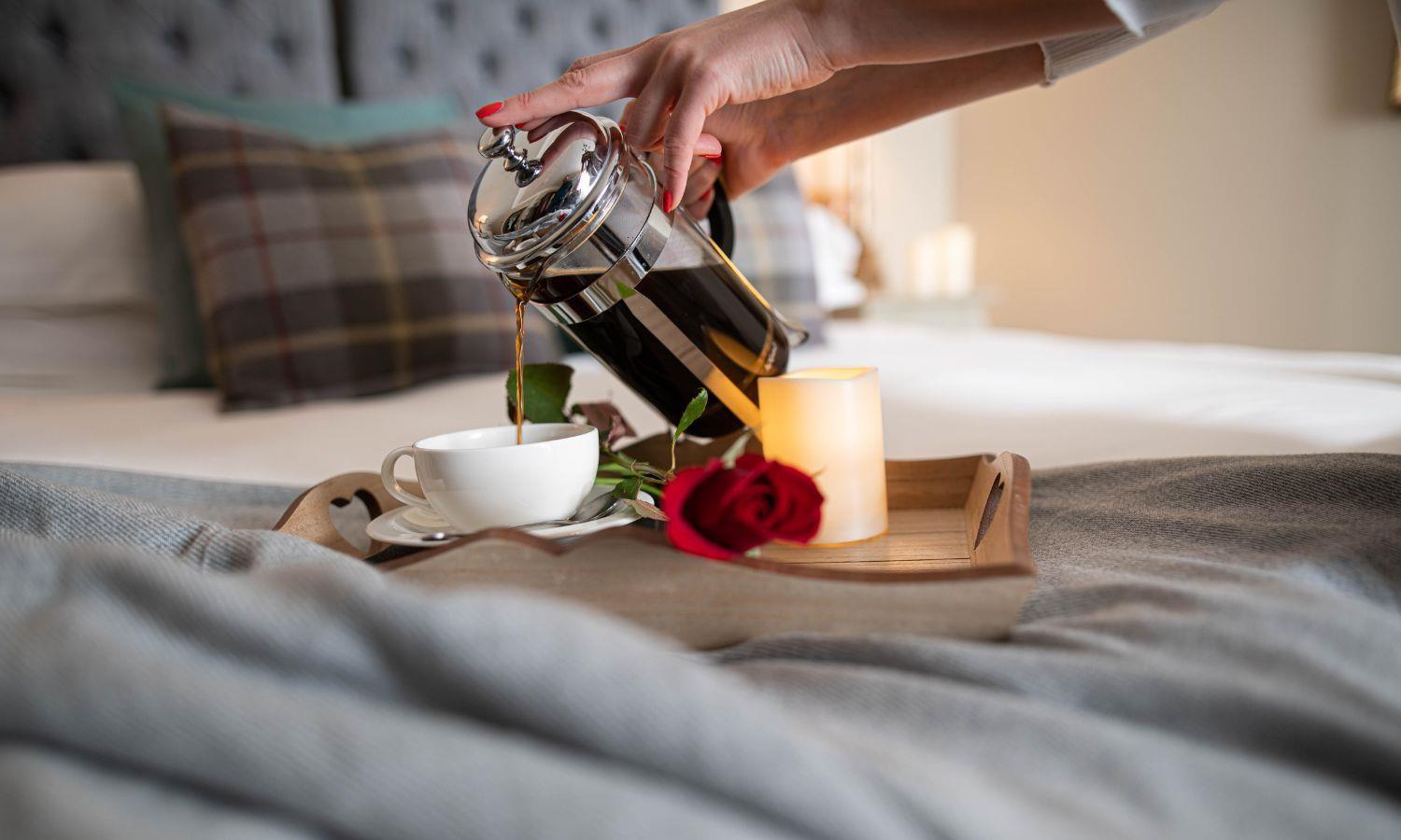 Woman's hand pouring coffee from cafetiere on bed tray with rose